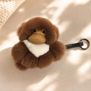 May include: A brown plush monkey keyring featuring a soft, fluffy texture, a smooth tan leather-like face with stitched detailing, and a white patch on the chest. The monkey is attached to a dark blue fabric strap and a silver-toned metal ring.