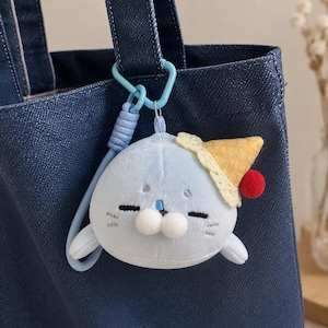Peut inclure: Un porte-clés en peluche bleu clair en forme de phoque, orné d'un petit chapeau en forme de cornet de glace jaune avec un pompon rouge. Le phoque a des yeux et des moustaches brodés, ainsi qu'un nez composé de deux sphères blanches.