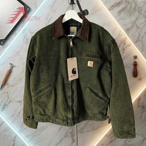 Op de afbeelding: Een donker olijfgroen canvas jack met een bruine corduroy kraag, een ritssluiting over de gehele lengte en twee schuine zakken aan de onderzijde. Een kleine borstzak met rits, een Carhartt-merklabel en een hangend papieren kaartje zijn zichtbaar.