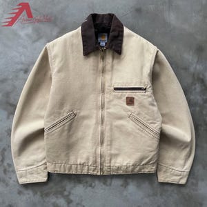 May include: A beige canvas jacket featuring a dark brown corduroy collar, a full-length front zipper, and long sleeves. The jacket includes two slanted lower pockets, a horizontal zippered chest pocket, and a small square brand patch on the front.