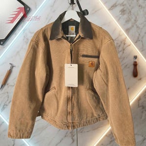Pode incluir: Uma jaqueta Carhartt bege com gola de veludo cotelê marrom escuro, zíper frontal completo e um bolso no peito com fechamento em zíper. A jaqueta inclui um logotipo quadrado da Carhartt, forro interno xadrez e uma etiqueta de papel branca.