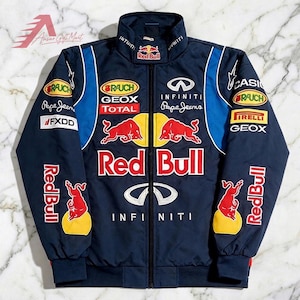 May include: A navy blue zip-up racing jacket featuring Red Bull branding, including red bull logos, yellow circles, and sponsor patches such as Infiniti, Rauch, Geox, Total, Pepe Jeans, FXDD, Casio, and Pirelli. The jacket has a high collar and long sleeves.