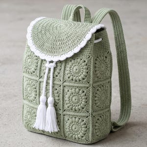 May include: A sage green crochet backpack constructed from square floral motifs. The bag features a rounded flap with a white scalloped edge, a white drawstring closure with two long white tassels, and two matching sage green shoulder straps.