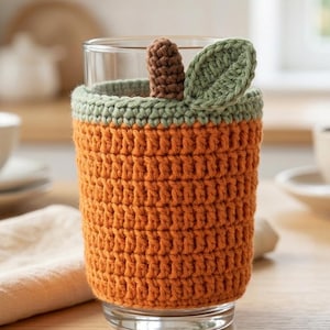 Peut inclure: Un verre transparent doté d'une housse en crochet orange texturée. La housse présente une bordure supérieure vert sauge, une petite tige en crochet marron et une feuille en crochet vert sauge fixée sur le bord supérieur.