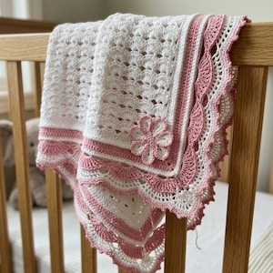 May include: A white crocheted blanket featuring a repeating shell stitch pattern, a dusty pink border with a scalloped edge, and a large, dimensional crocheted flower appliqué attached to one corner of the fabric, creating a textured appearance.