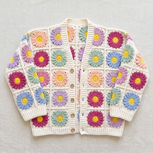 May include: A long-sleeve cardigan made of cream-colored yarn, featuring a repeating pattern of crocheted flower squares in shades of pink, purple, blue, and peach, each with a yellow center, finished with a row of five brown buttons down the front.