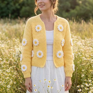 May include: A bright yellow long-sleeve crochet cardigan featuring a repeating pattern of white and yellow daisy flower motifs. The open-front garment has a ribbed hem and cuffs, worn over a simple white top and a white skirt.