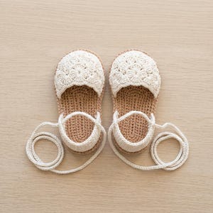 Può includere: Un paio di sandali lavorati all'uncinetto a mano con un motivo floreale color crema sulla punta e una suola color marrone chiaro. I sandali includono lunghi lacci all'uncinetto color crema da avvolgere attorno alla caviglia.