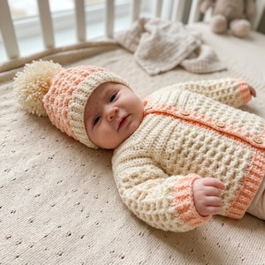 May include: A crochet baby set consisting of a long-sleeved cardigan and a matching beanie. The cardigan is cream-coloured with peach-coloured ribbed cuffs and trim, featuring wooden buttons. The beanie is peach-coloured with a cream ribbed brim and a large cream pom-pom.