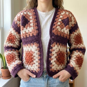 May include: A long-sleeve open-front cardigan constructed from crocheted granny squares in a repeating pattern of deep purple, burnt orange, and cream yarn. The garment features a V-neckline and a cropped silhouette with ribbed cuffs.
