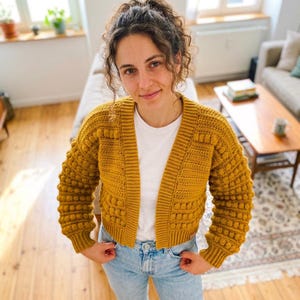 May include: A mustard yellow crochet cardigan featuring a textured bobble stitch pattern on the sleeves and body, a ribbed V-neckline, and ribbed cuffs. The garment is worn over a plain white t-shirt and light blue denim jeans.