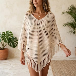 May include: A cream-colored crochet poncho featuring an open diamond-shaped knit pattern, a V-neckline, and long fringe detailing along the hem. The garment has a loose, flowing silhouette with wide sleeves and a pointed front hemline.