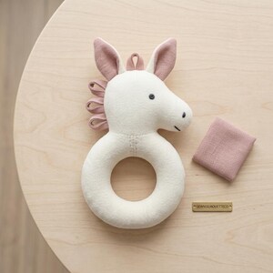 May include: A cream-colored fabric animal rattle shaped like a horse head with a circular base, featuring dusty rose-colored ears and a mane made of fabric loops. A small square dusty rose fabric swatch and a gold tag labeled SEWNSILHOUETTECO.