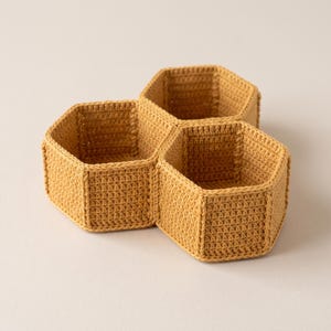 May include: A mustard yellow crochet basket featuring a honeycomb design with three interconnected hexagonal compartments. The structure is made of dense, uniform yarn stitches, creating a sturdy, open-top container with defined vertical walls.