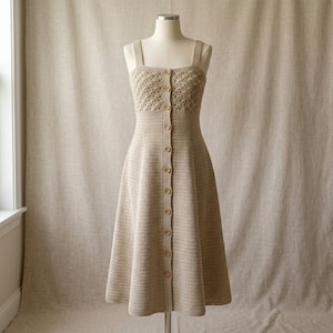 Puede incluir: Un vestido beige sin mangas con un corpiño de ganchillo con patrón de diamantes y una falda de ganchillo lisa. El vestido tiene un escote cuadrado, tirantes anchos y una fila vertical de diez pequeños botones redondos de madera al frente.