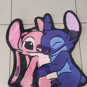 May include: A plush rug shaped like the characters Stitch and Angel hugging. The rug features a dark purple Stitch and a light pink Angel with large ears, black eyes, and a thick black border outlining the entire design against a tiled floor.