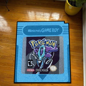 May include: A rectangular tufted rug shaped like a Nintendo Game Boy video game cartridge. The rug is light blue with the text Nintendo GAME BOY at the top. The center features the Pokémon Crystal Version game label with the character Suicune.