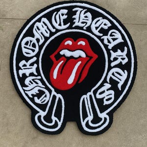 Puede incluir: Una alfombra circular con mechones que presenta un fondo negro con un gráfico central rojo y blanco de una boca con la lengua afuera. El texto CHROME HEARTS está bordado en letras góticas blancas alrededor del borde circular de la alfombra.