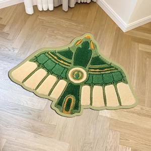 May include: A tufted rug shaped like a stylised bird with wings spread wide. The design features deep green, light green, cream, and gold-coloured yarn. The central body has a circular emblem, and the wing feathers are segmented in cream and green.
