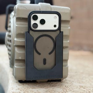 May include: A black plastic phone mount attached to a tan, rectangular, ribbed speaker. The mount features a circular magnetic ring design and the embossed text Jace Ketton. A smartphone with a triple-lens camera is secured within the mount.