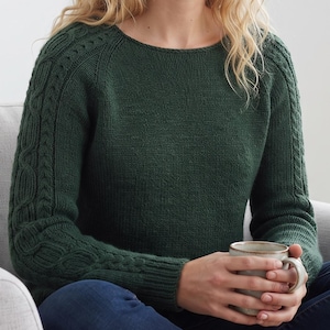 Può includere: Un maglione a maniche lunghe verde bosco scuro con scollo rotondo e intricati motivi a trecce lungo le maniche. Il capo presenta una lavorazione a maglia a coste sui polsini e un corpo in maglia liscia.