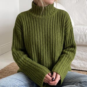 May include: An olive green jumper featuring a high mock neck collar and a vertical ribbed knit texture. The garment has long sleeves with a relaxed fit and a straight hemline, crafted from a knit fabric that provides a consistent vertical pattern.