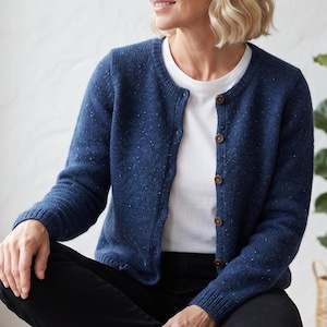 May include: A navy blue speckled knit cardigan featuring a round neckline, long sleeves with ribbed cuffs, and a front button closure with five dark brown buttons. The cardigan is worn over a plain white t-shirt and paired with black trousers.