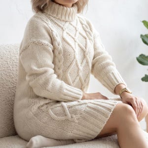 May include: An off-white, long-sleeve jumper dress featuring a chunky knit texture, a high turtleneck collar, and a prominent cable knit pattern running vertically down the front. The garment has ribbed cuffs and a ribbed hemline at the bottom.