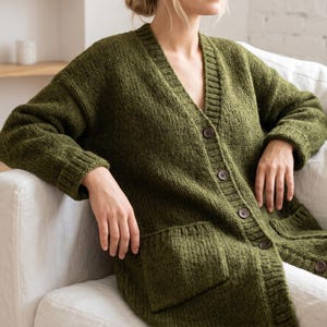 Può includere: Un cardigan in maglia verde oliva con scollo a V, maniche lunghe con polsini a coste e chiusura frontale con bottoni. Il capo presenta due ampie tasche applicate sulla parte inferiore, realizzate in filato spesso e testurizzato.