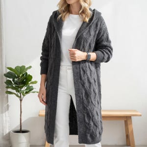Peut inclure: Un long cardigan à capuche gris foncé ouvert sur le devant, doté d'un motif en tricot torsadé épais sur les panneaux avant et les manches. Le vêtement présente des manches longues avec des poignets et un ourlet côtelés, porté sur un t-shirt blanc.