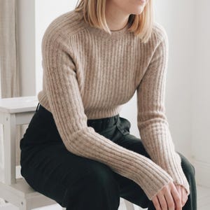 Op de afbeelding: Een beige trui met lange mouwen, een verticaal geribbelde textuur en een ronde halslijn, gecombineerd met een effen zwarte broek. De trui heeft een aansluitend silhouet met raglanmouwen en is gemaakt van een zachte, gebreide stof.