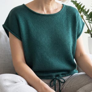 May include: A dark emerald green knit top featuring a wide boat neckline, short cap sleeves with rolled edges, and a drawstring hemline that allows for a cinched fit. The garment is crafted from a solid-colored, textured knit fabric.