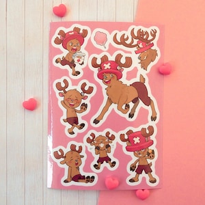 Puede incluir: Una hoja de pegatinas rosa con múltiples ilustraciones del personaje Tony Tony Chopper de One Piece. Las pegatinas muestran al reno de pelaje marrón en varias poses, usando un sombrero rojo con una cruz blanca, e incluyen un icono de algodón de azúcar.