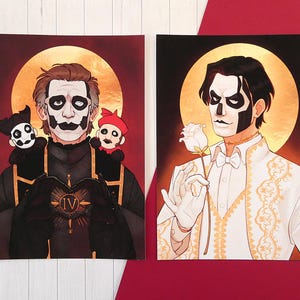 May include: Two rectangular art prints featuring stylized illustrations of figures with face paint. The left print shows a figure in black robes with two small puppets and a heart-shaped IV symbol. The right print shows a figure in a white suit holding a white rose.