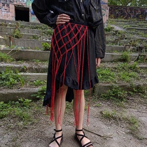 May include: A black long-sleeved button-down shirt with thin vertical stripes paired with a black skirt featuring a ruffled hem. A red macrame net overlay with long hanging fringe cords is worn over the skirt. Black strappy sandals are worn.