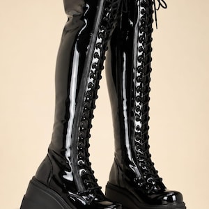 May include: A pair of black patent leather knee-high boots featuring a full-length front lace-up closure with black laces, a side zipper, and a thick, chunky black platform lug sole with a wedge heel design for added height and stability.