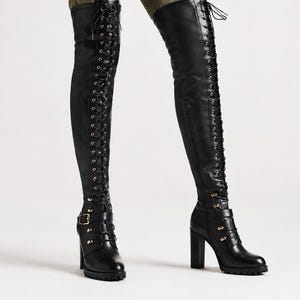 May include: Black over-the-knee boots featuring a full-length front lace-up design with metal eyelets, a side zipper, a buckled strap detail at the ankle, a high block heel, and a lugged sole. The boots extend above the knee in a smooth finish.