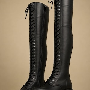 May include: A pair of black knee-high boots featuring a full-length lace-up front with metal eyelets, black laces tied at the top, a side zipper closure, and a thick, chunky lug sole with a block heel. The boots have a smooth, matte finish.