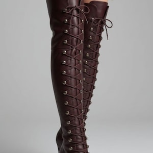 May include: Dark burgundy over-the-knee boots featuring a full-length front lace-up closure with thin matching laces and metallic eyelets. The boots have a rounded toe, a high stiletto heel, and a smooth finish that extends above the knee.