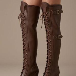 May include: A pair of dark brown over-the-knee boots featuring a full-length front lace-up design with matching brown laces, a block heel, a platform sole, and two adjustable straps with bronze-toned metal buckles on the outer side of each shaft.