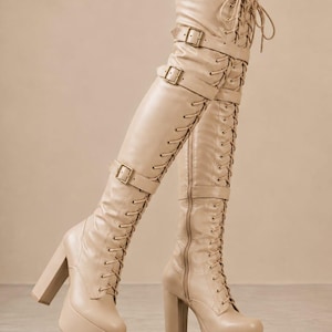 May include: Beige over-the-knee boots featuring a high block heel, a thick platform sole, and a full-length lace-up front. The boots include three horizontal straps with metallic buckles across the shaft and a side zipper for closure.