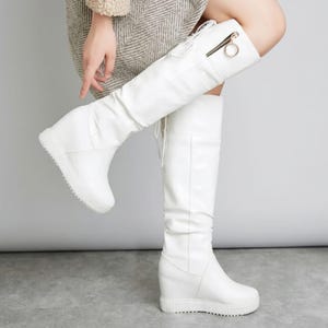 May include: A pair of white knee-high boots featuring a hidden wedge heel, a textured rubber sole, and a decorative side zipper with a circular gold-tone pull. The boots have a smooth finish and include lace-up detailing at the back of the shaft.