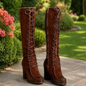 May include: A pair of knee-high brown suede boots featuring a full-length front lace-up design with matching brown laces, a block heel, and a side zip closure. The boots have a rounded toe and visible stitching along the front panel edges.