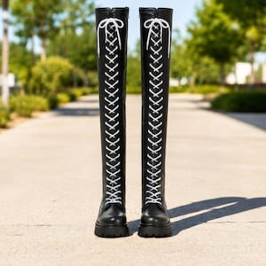 May include: A pair of black over-the-knee boots featuring a full-length front lace-up design with white laces tied in a bow at the top. The boots have a rounded toe, a thick lug sole, and a smooth finish, extending well above the knee.