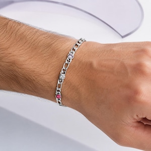 May include: A silver-toned curb chain bracelet featuring three circular bezel-set stones in shades of light purple, clear white, and vibrant pink, arranged linearly along the metallic links of the wrist accessory.