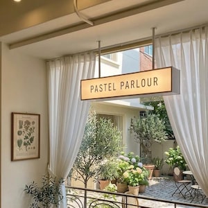 May include: A rectangular, illuminated hanging sign with the text PASTEL PARLOUR in black letters. The sign is suspended from the ceiling above a balcony with a metal railing, overlooking a courtyard filled with potted plants and trees.