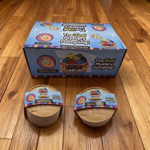 May include: A rectangular blue cardboard box labelled The Viral Mystery Dumpling with cartoon illustrations, accompanied by two round tan plastic containers, each wrapped in a paper band featuring the same product branding and logo.