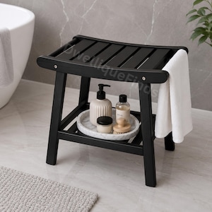 May include: A black slatted shower stool with four legs and a lower shelf. A white towel hangs from the side. On the shelf sits a round white marble tray holding a beige soap dispenser, a small jar with a black lid, and a wooden scrub brush.