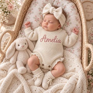 Könnte beinhalten: Ein cremefarbener, gestrickter Langarm-Strampler mit dem in rosa Schreibschrift aufgestickten Namen Amelia auf der Brust, dazu ein passendes geripptes Stirnband mit großer Schleife, cremefarbene Strickschühchen mit kleinen Schleifen und ein Plüschhase.