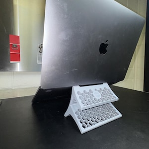 May include: A white laptop stand featuring a two-tiered structure with a repeating hexagonal honeycomb pattern. The stand is positioned underneath a dark gray laptop, providing an angled elevation for the device while resting on a flat surface.
